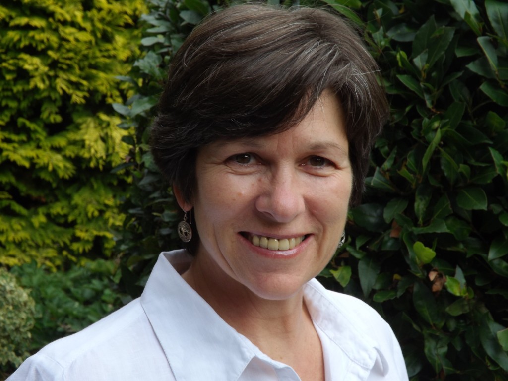 Sue Chappell, smiling at the camera in a white shirt, standing in front of a dark hedge with a golden conifer behind her.